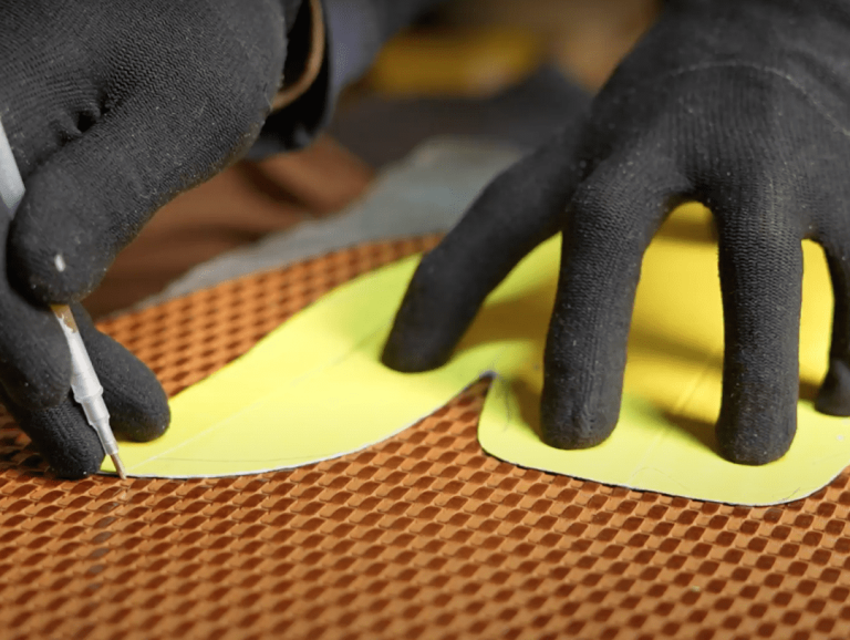 A craftsman wearing gloves carefully cutting a shoe upper pattern from a yellow template.